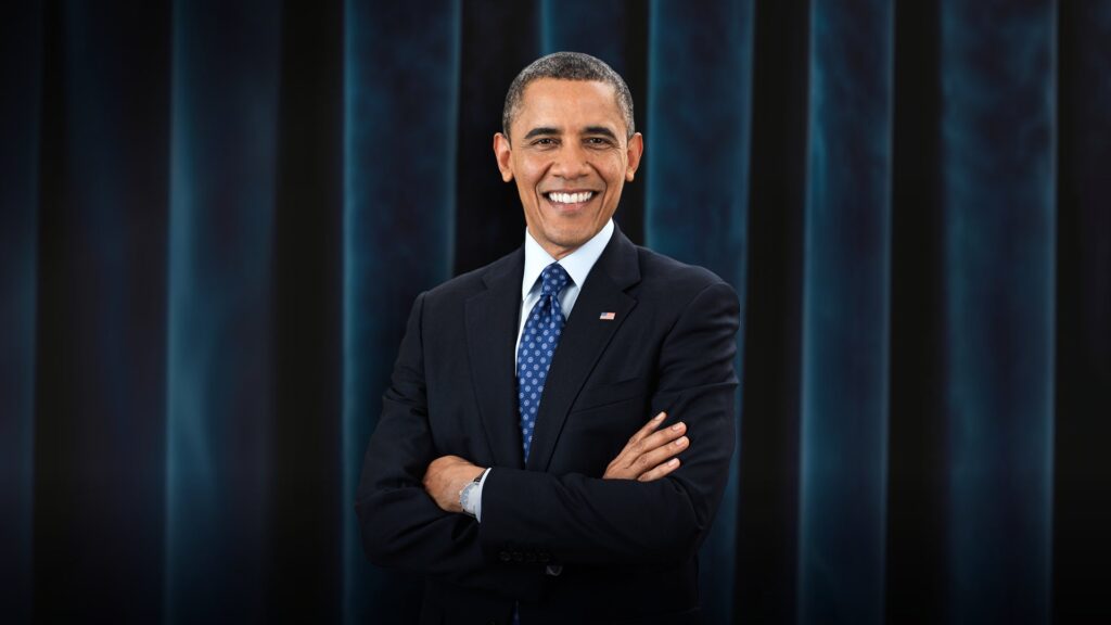 A formal headshot of former U.S. President Barack Obama, smiling warmly and wearing a classic dark suit and blue tie.