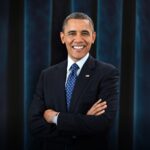 A formal headshot of former U.S. President Barack Obama, smiling warmly and wearing a classic dark suit and blue tie.