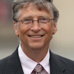 Bill Gates smiling, wearing a black suit, a tie, and sunglasses.
