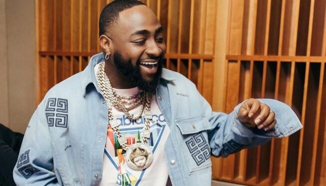 A close-up portrait of Nigerian Afrobeats artist David Adeleke, smiling and showing his dimples, styled in fashionable streetwear and diamond jewelry.