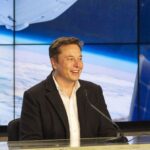 Elon Musk speaking at a tech conference, gesturing with his hands while standing on a stage with a SpaceX rocket graphic on the screen behind him.