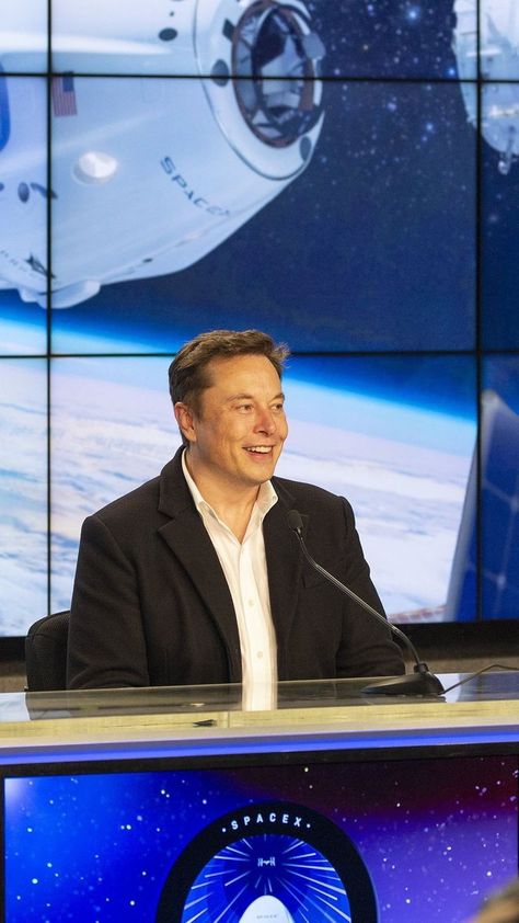 Elon Musk speaking at a tech conference, gesturing with his hands while standing on a stage with a SpaceX rocket graphic on the screen behind him.