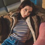 A high-fashion, minimalist portrait of internet personality Emma Chamberlain, styled in chic retro-inspired clothing with her signature deadpan, artistic gaze.