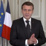 An official portrait of French President Emmanuel Macron, standing confidently in a dark suit next to the French flag.