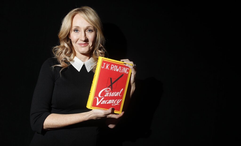 A portrait of author Joanne Rowling smiling and holding up a copy of her new book at a book launch event