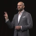 A clean, professional photo of author James Clear, wearing a tailored suit against a minimalist background while speaking in a conference.