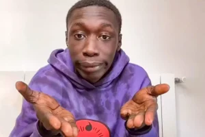 A playful portrait of content creator Khaby Lame, making his famous hand-shrugging gesture with an expressive, deadpan facial expression.