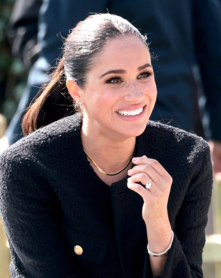 A beautiful, professional headshot of Meghan, Duchess of Sussex, smiling warmly with her hair pulled back, wearing clean, minimalist attire.