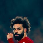 A close-up of footballer Mohamed Salah on the pitch, sporting his signature curls and a focused expression.