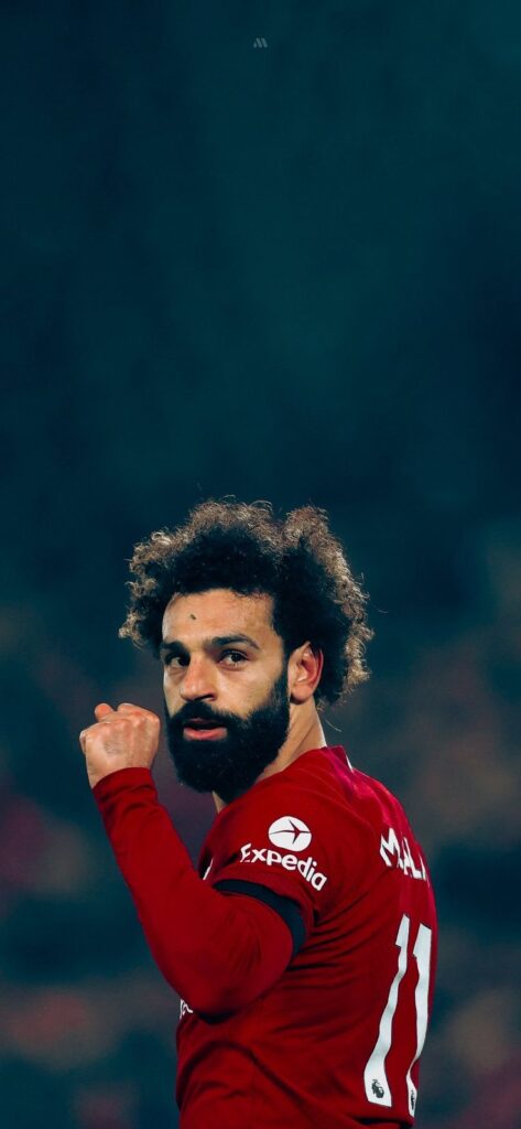 A close-up of footballer Mohamed Salah on the pitch, sporting his signature curls and a focused expression.