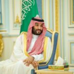 Mohammed bin Salman, Crown Prince of Saudi Arabia, wearing traditional Saudi thobe and ghutra and sitting behind the flag.