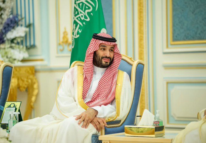 Mohammed bin Salman, Crown Prince of Saudi Arabia, wearing traditional Saudi thobe and ghutra and sitting behind the flag.