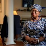 A close-up portrait of WTO Director-General Dr. Ngozi Okonjo-Iweala, smiling warmly in her signature vibrant Ankara print dress and traditional headtie (gele).