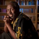 Ngũgĩ wa Thiong'o, wearing a traditional embroidered African tunic sitting behind a book shelf, smiling warmly.