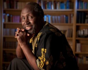 Ngũgĩ wa Thiong'o, wearing a traditional embroidered African tunic sitting behind a book shelf, smiling warmly.