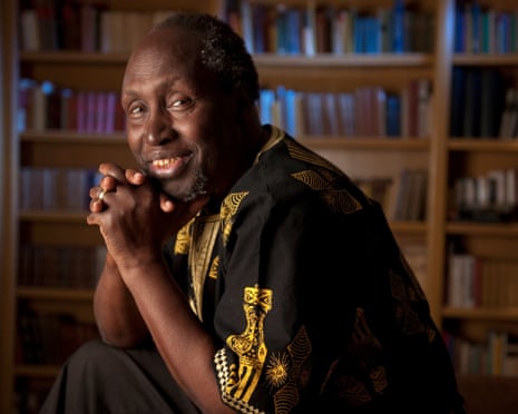 Ngũgĩ wa Thiong'o, wearing a traditional embroidered African tunic sitting behind a book shelf, smiling warmly.