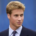A formal portrait of Prince William, the Prince of Wales, smiling in a tailored dark suit and tie against a neutral background.