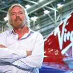 A smiling portrait of Virgin Group founder Richard Branson, showcasing his signature blonde hair, beard, and a casual open-collar white shirt.