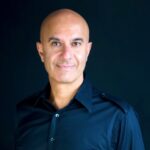 A portrait of leadership expert and author Robin Sharma, smiling confidently in a sharp, casual navy blue T-shirt in a neutral background.