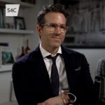A professional portrait of actor Ryan Reynolds smiling, wearing a classic suit and glasses.
