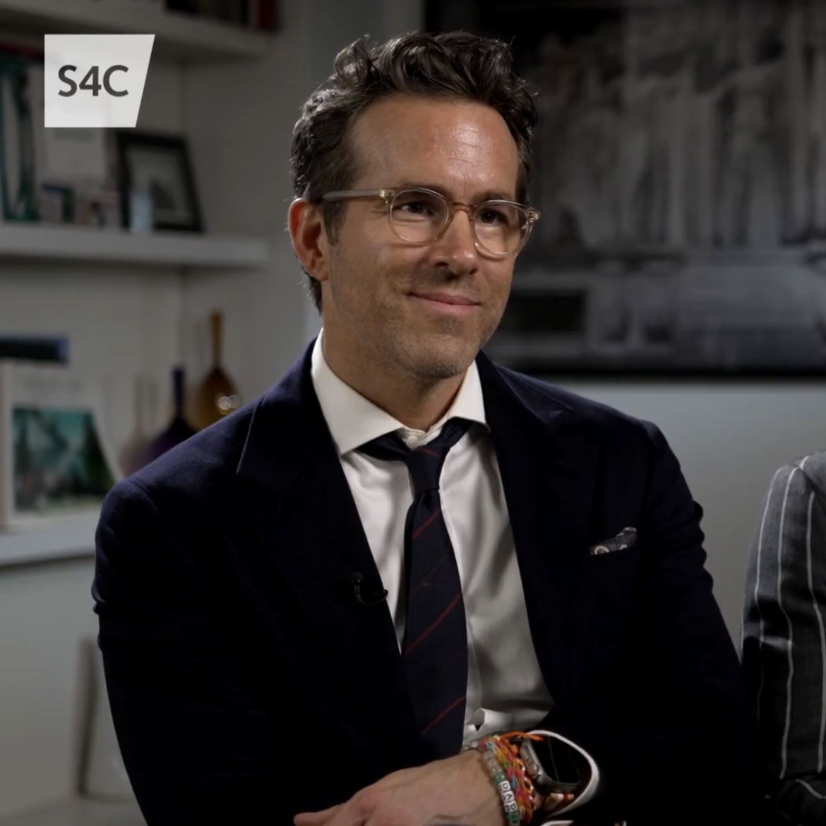 A professional portrait of actor Ryan Reynolds smiling, wearing a classic suit and glasses.