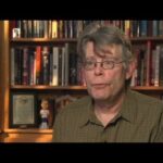 A candid portrait of author Stephen King sitting behind a well-stocked library bookshelf.