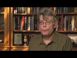 A candid portrait of author Stephen King sitting behind a well-stocked library bookshelf.