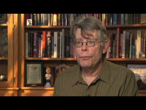 A candid portrait of author Stephen King sitting behind a well-stocked library bookshelf.