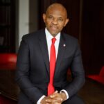 A professional portrait of Nigerian economist and philanthropist Tony Elumelu, wearing a sharp tailored suit with his signature red tie, smiling warmly.