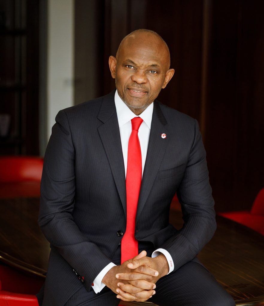 A professional portrait of Nigerian economist and philanthropist Tony Elumelu, wearing a sharp tailored suit with his signature red tie, smiling warmly.