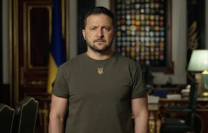 A close-up portrait of Ukrainian President Volodymyr Zelenskyy, looking serious and resolute in his signature olive-green military tee.