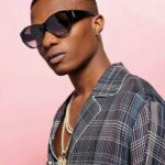 A stylish, high-fashion portrait of Nigerian musician Wizkid, wearing designer sunglasses and trendy minimalist apparel.