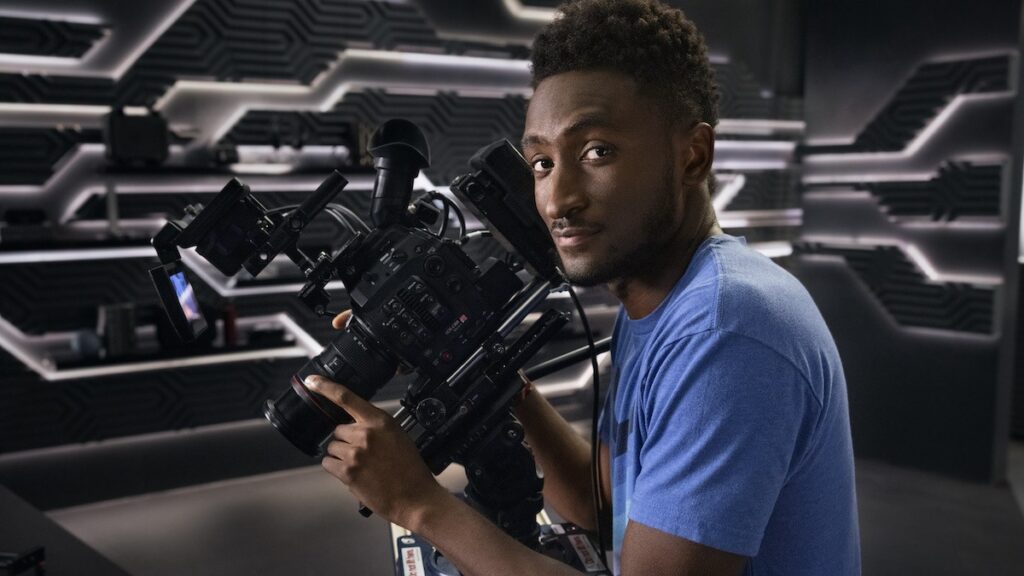 A crisp professional portrait of tech reviewer Marques Brownlee, smiling in a casual blue t-shirt against a studio background.